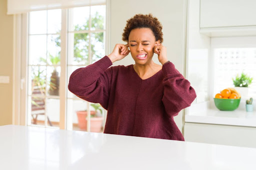 A woman sitting in a kitchen and covering her ears because of a loud noise.