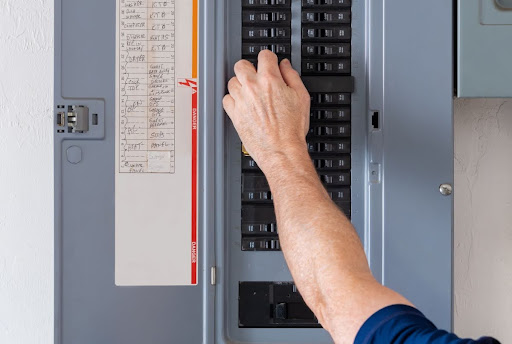 A man's hand reaching up to reset a tripped circuit breaker on an electrical panel.
