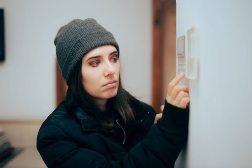 A woman in a winter hat and coat adusting a thermostat in a home.