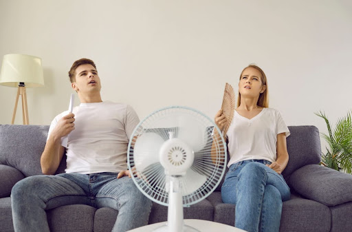 A man and woman sitting on a couch fanning themselves while a fan blows on them.