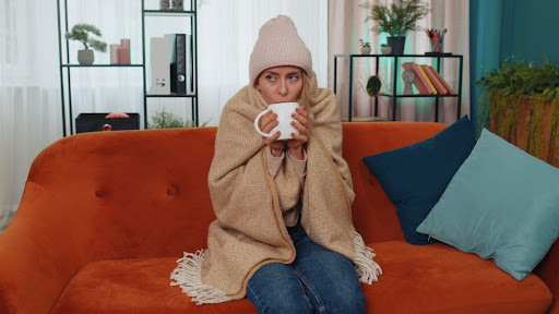 A woman with a winter hat and blanket wrapped around her shoulders sitting on a couch with a mug.